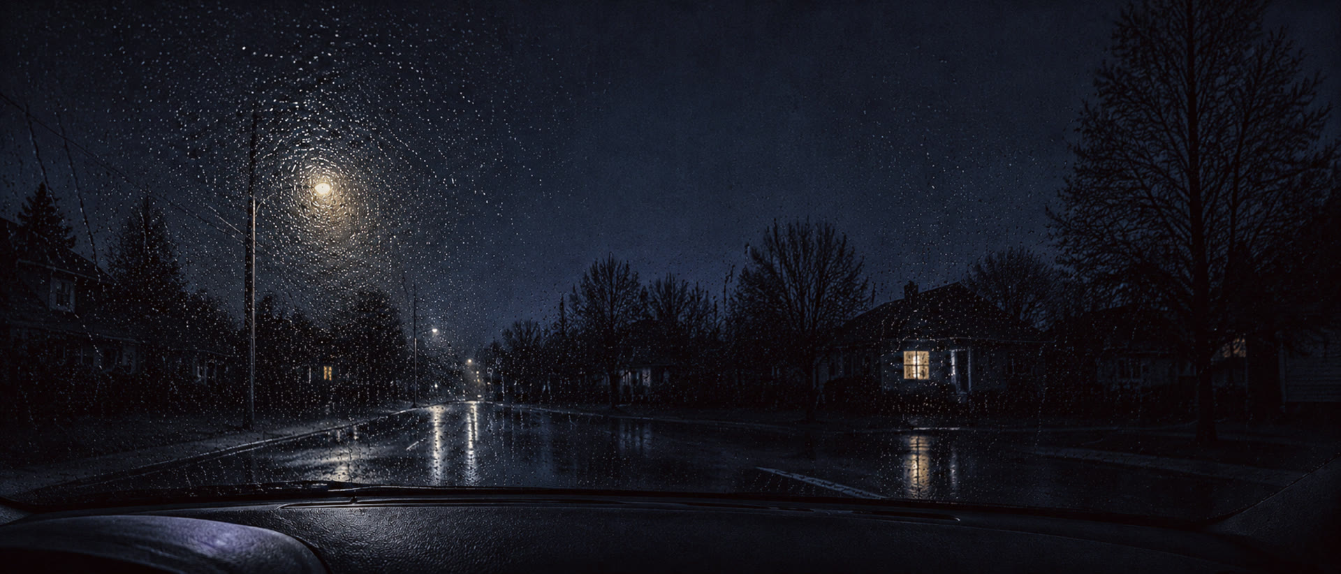 A rain speckled windshield at night, one warm lit window glowing in the dark